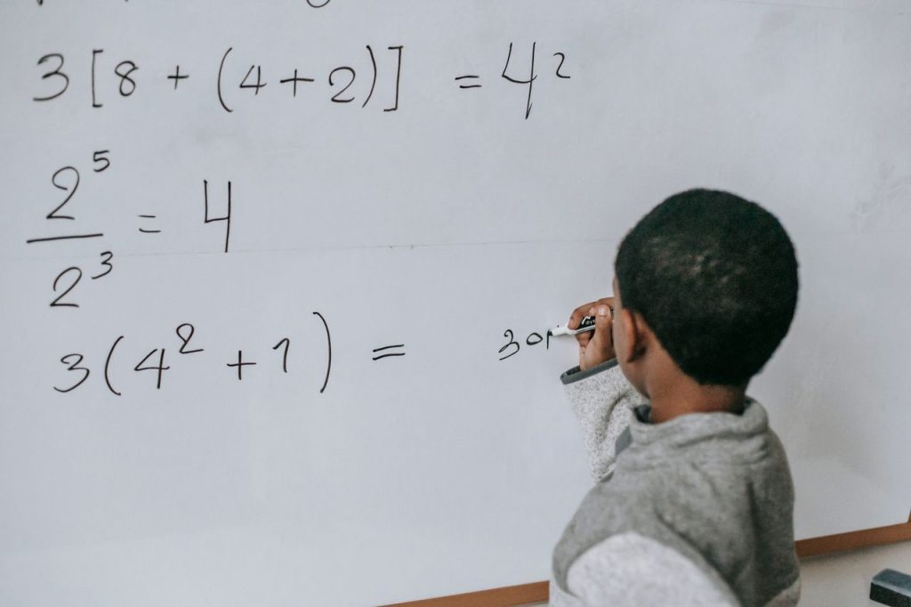 Back view of anonymous African American boy writing on whiteboard while doing sums on mathematics class